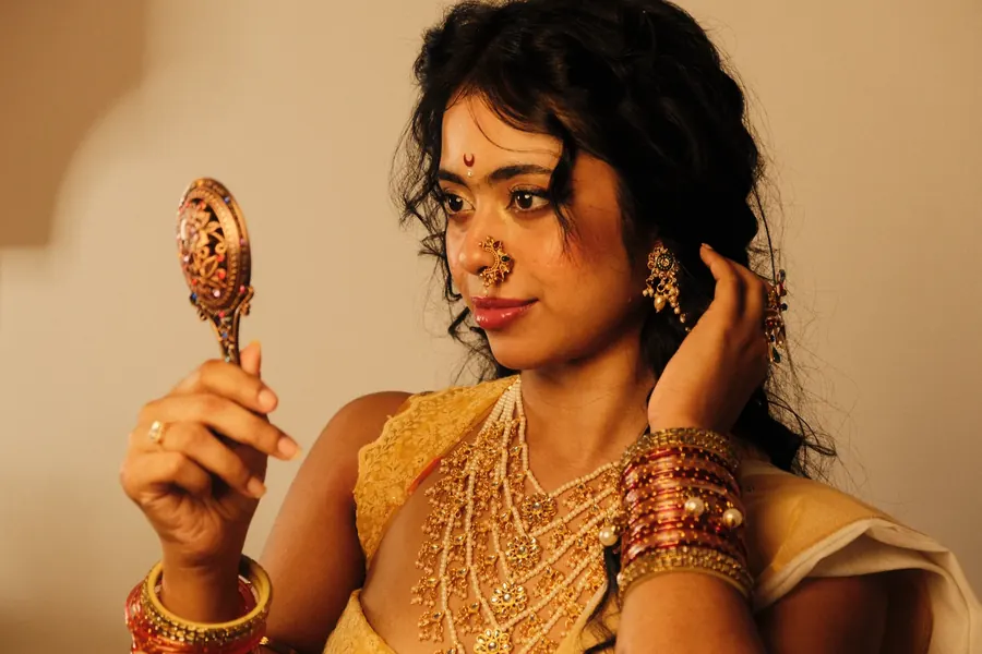 Sandipta in traditional jewellery and a bindi, holding an ornate hand mirror