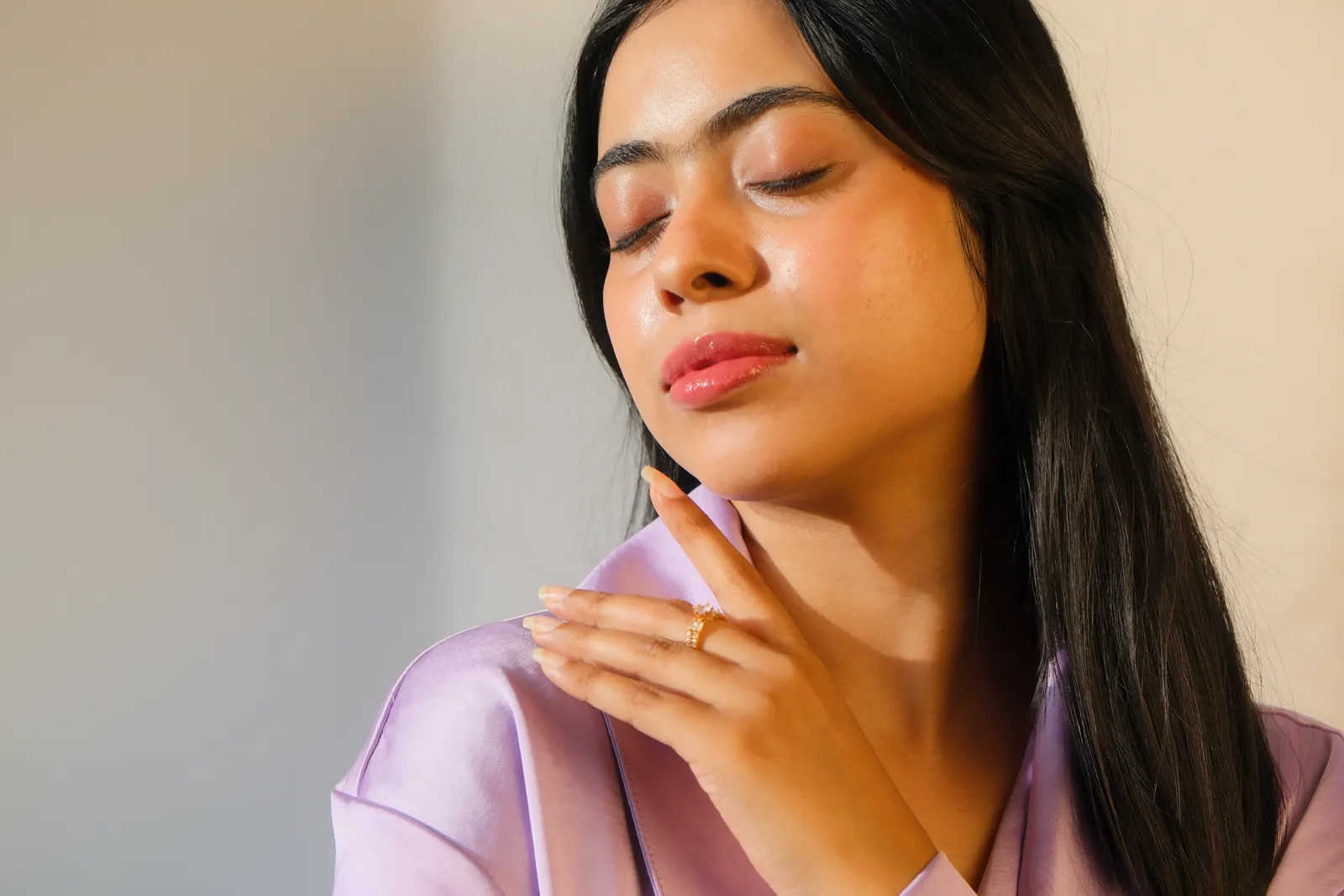 Sandipta in warm side light, eyes closed, one hand at her neck, wearing a lilac robe